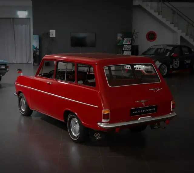 Röd veteranbil med ålder, en Opel Caravan, står i en bilhall bredvid en blå bil och en tävlingsbil.