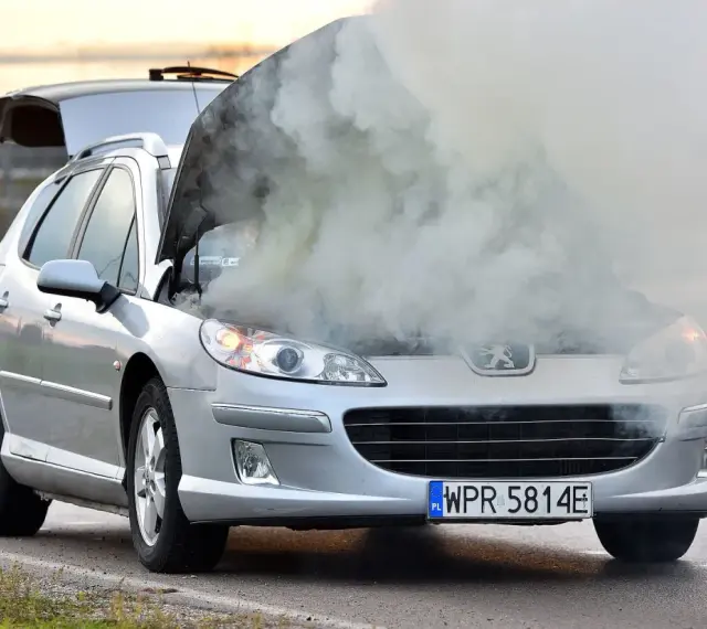 Srebrny samochód Peugeot z otwartą maską, z której wydobywa się gęsty dym.