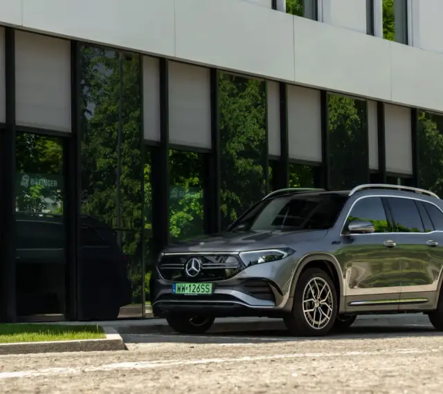 Nowy Mercedes 4x4, elegancki SUV, zaparkowany przed nowoczesnym budynkiem z zielonymi odbiciami drzew w szybach.