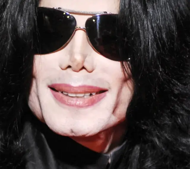 Michael Jackson w okularach przeciwsłonecznych i czarnych włosach. Jego uśmiech i twarz budzą pytania, jak zmarł Michael Jackson.
