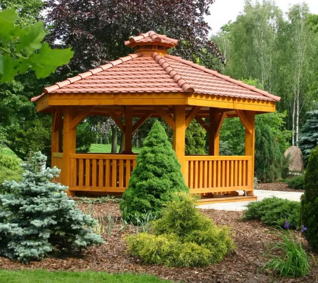 Drewniana altana z czerwonym, ceramicznym dachem, otoczona bujną zielenią. Idealne miejsce na relaks, a dach z dachówki to świetny pomysł, czym pokryć dach altany tanio i estetycznie.