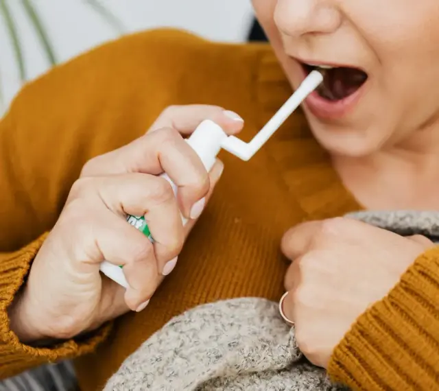 Kobieta aplikuje spray do gardła, by złagodzić ból i pieczenie spowodowane przez afty w gardle.