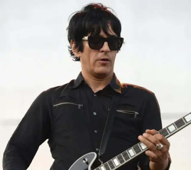 Un des membres de Eagles of Death Metal, guitariste en chemise noire et lunettes de soleil, joue sur scène.