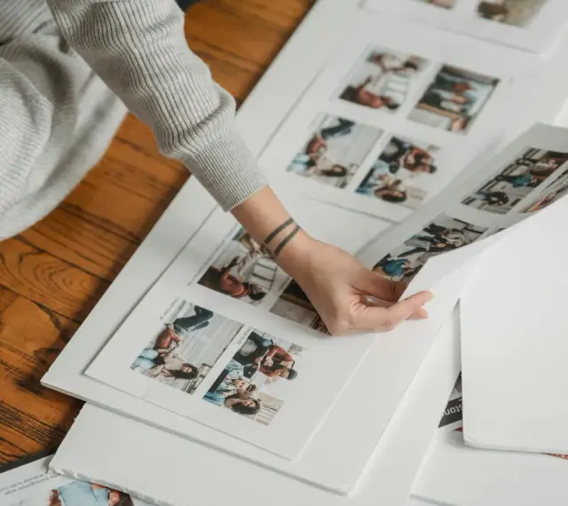Osoba przegląda wydruki zdjęć, planując, jak zrobić fotoksiążkę. Na papierze widać małe zdjęcia ludzi.
