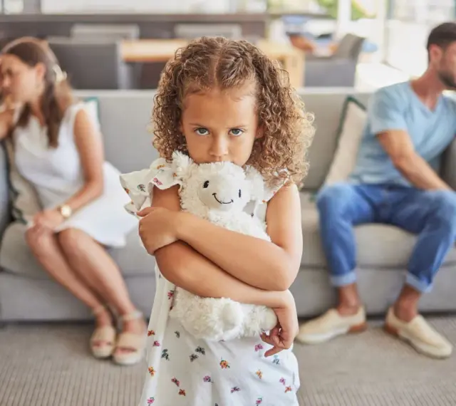 Ein Kind umklammert sein Stofftier, während die Eltern im Hintergrund getrennt sitzen. Ein Bild, das die emotionale Belastung eines Trennungsjahres mit Kindern widerspiegelt.