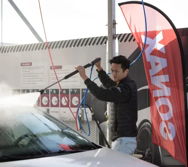 Man using a pressure washer on a car windshield, with a SONAX banner in the background.