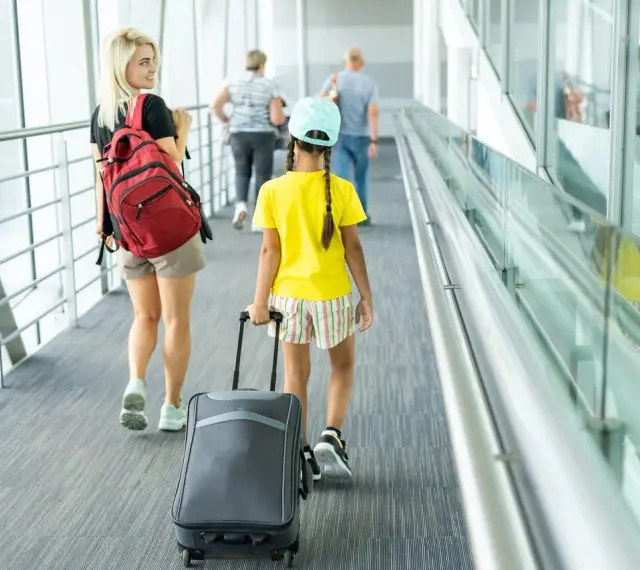 Młoda kobieta z plecakiem i dziewczynka z walizką idą korytarzem lotniska. To może być ich wyjazd nieletniego za granicę bez rodziców.