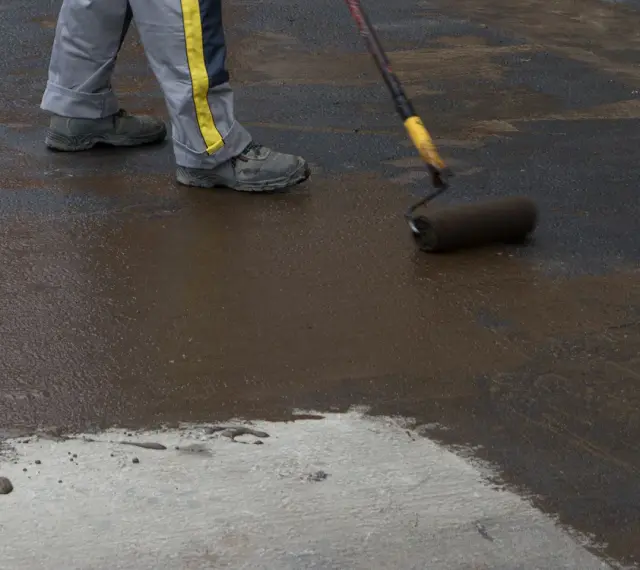Pracownik wykonuje hydroizolację chudziaka, nakładając masę bitumiczną wałkiem.