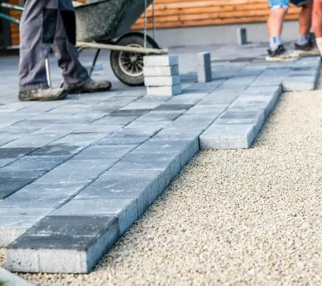 Układanie płyt betonowych na podjeździe – alternatywa dla kostki. Pracownicy budowlani układają szare płyty na podsypce żwirowej.