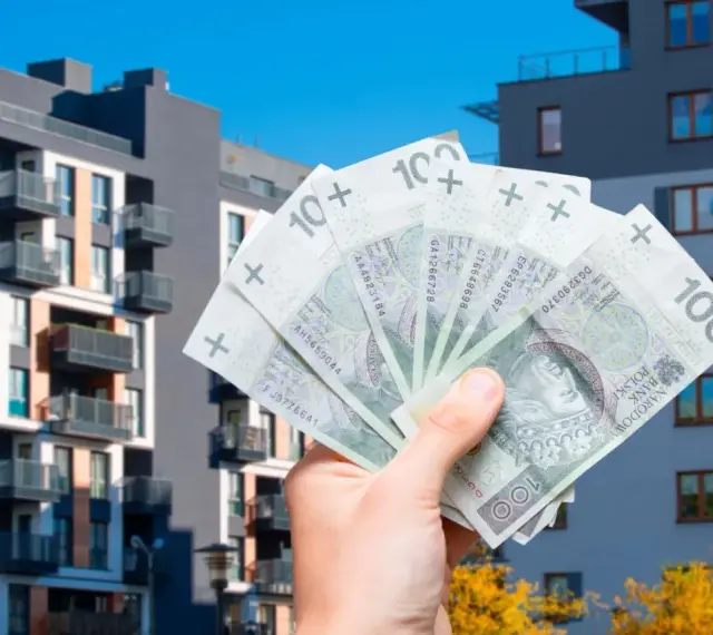 Dłoń trzyma banknoty 100 zł na tle nowoczesnych budynków mieszkalnych. Symbolizuje to podatek od sprzedaży nieruchomości.