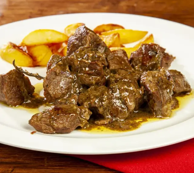 Un saporito abbacchio alla cacciatora, tenero e succulento, servito con patate arrosto su un piatto bianco.