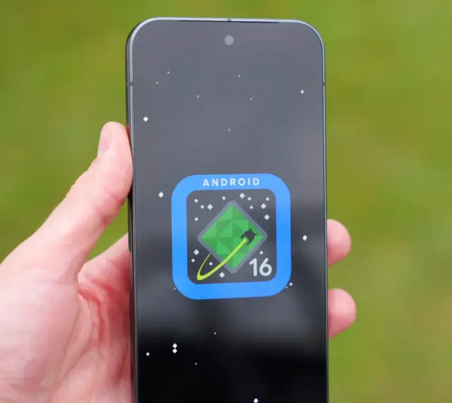 Dłoń trzyma smartfon z ikoną Android 16 na ekranie, przypominającą zielony kryształ na tle kosmosu.
