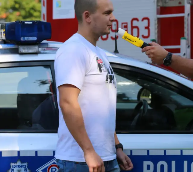 Policjant przeprowadza procedurę badania alkomatem. Mężczyzna dmucha w urządzenie przy radiowozie.