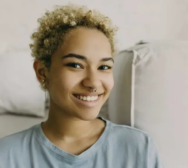 Uśmiechnięta młoda kobieta z kręconymi blond włosami i kolczykiem w nosie (septum piercing).