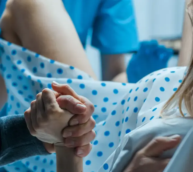 Wsparcie bliskiej osoby podczas porodu. Dłoń partnera trzyma dłoń rodzącej, która ma już widoczny czop śluzowy.