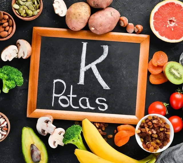 Tablica z napisem "K Potas" otoczona owocami i warzywami bogatymi w potas, jak banany, awokado, pomidory, ziemniaki, brokuły, rodzynki, morele, kiwi, migdały, pestki dyni. Pomaga zapobiegać hipokaliemii.