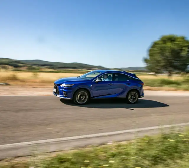 Niebieski Lexus 450h pędzi po wiejskiej drodze, otoczony zielenią i wzgórzami.