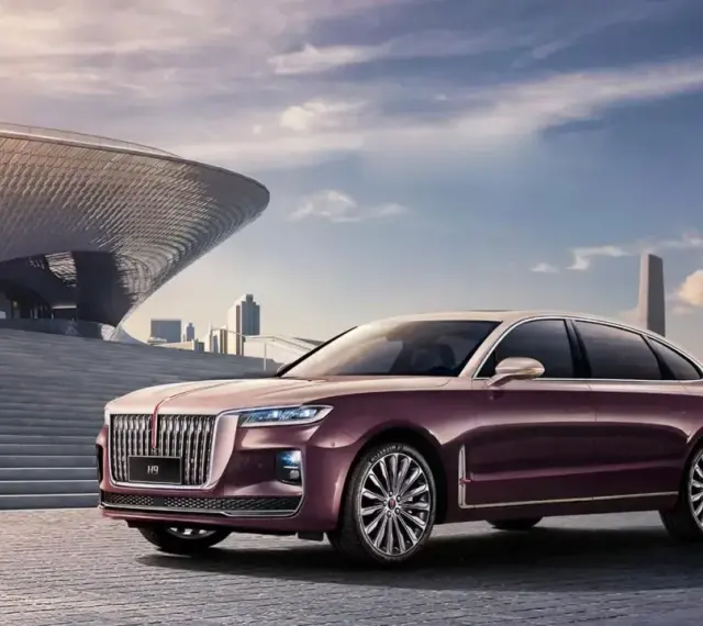 Elegancki, bordowy Hongqi H9 z chromowanymi detalami stoi przed nowoczesnym budynkiem o falistej fasadzie, w tle miejski krajobraz.