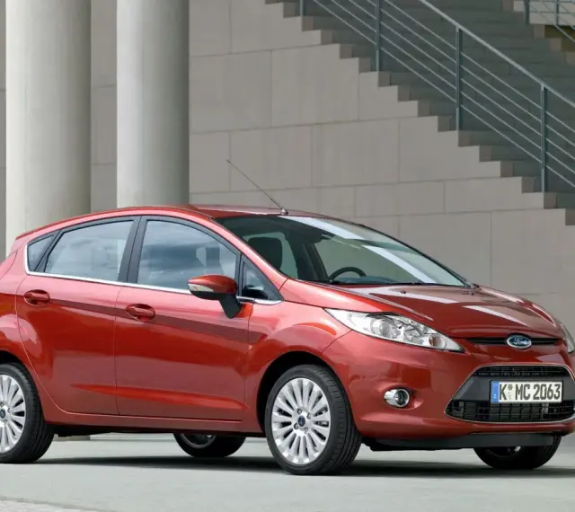 Czerwony Ford Fiesta MK6 zaparkowany przed nowoczesnym budynkiem z betonowymi kolumnami i schodami.