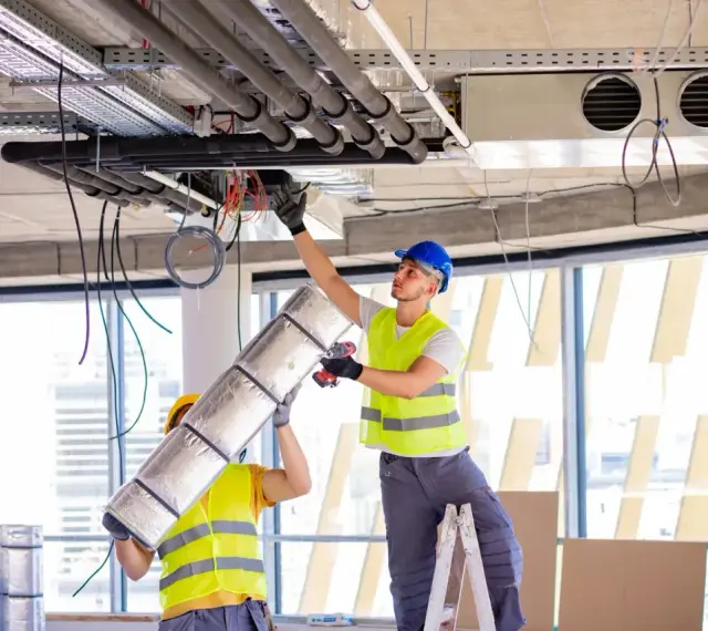 Dwóch pracowników montuje systemy HVAC, instalując izolację na rurach. To kluczowy element zapewniający komfort cieplny.