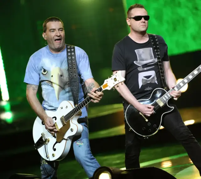 Dwóch członków Kobranocki gra na gitarach podczas koncertu. Jeden w koszulce z orłem, drugi w czarnej z cylindrem i muszką.