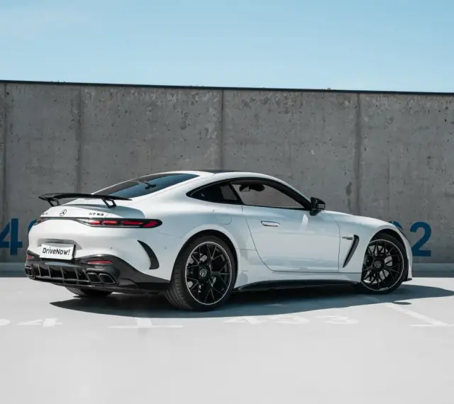 Biały Mercedes GT 63 z czarnym spojlerem i felgami, zaparkowany pod betonową ścianą z niebieskimi cyframi.