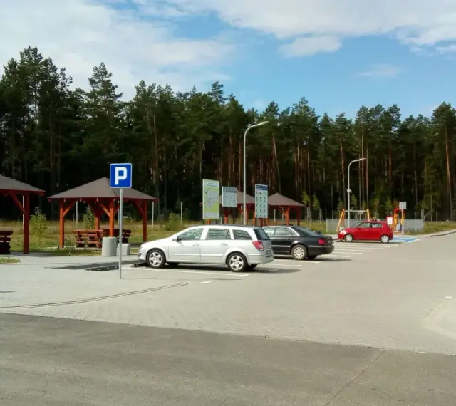 Parking przy lesie, z wiatami i ławkami. W tle las, a na parkingu kilka samochodów. To idealne miejsce na postój w drodze, może nawet w pobliżu **Orlen Sople**.