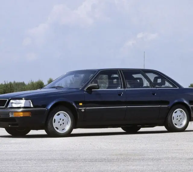 Ciemnoniebieski Audi V8 z lat 90. stoi na pustej drodze, z drzewami w tle.