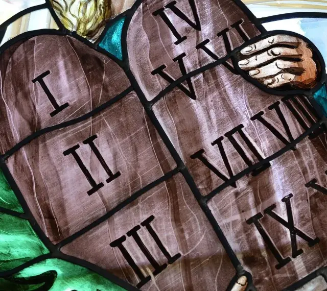 Witraż przedstawia kamienne tablice z wyrytymi cyframi rzymskimi, symbolizujące dekalog przykazania. Widoczne są cyfry I, II, III, IV, V, VI, VII, VIII, IX, X.
