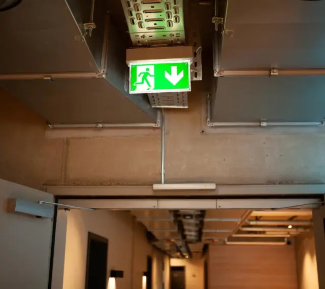 Zielone światła awaryjne wskazują drogę ewakuacji w korytarzu z widocznymi instalacjami wentylacyjnymi i betonowymi ścianami.
