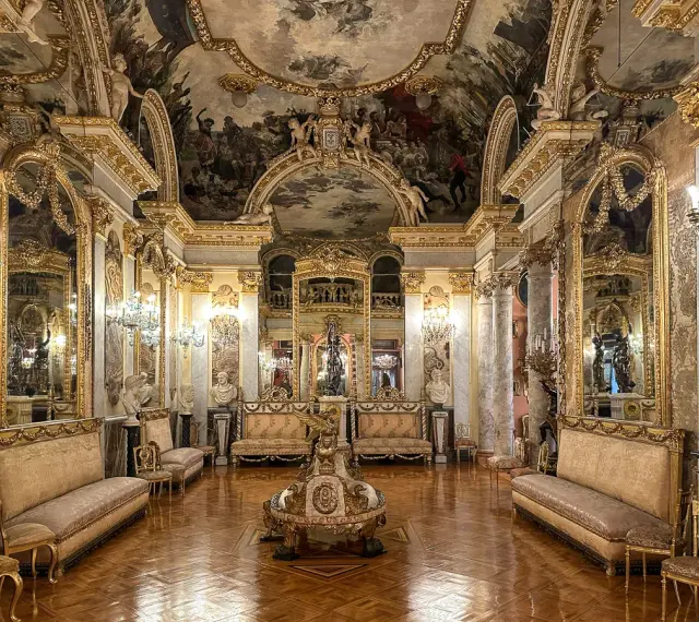 Złocone wnętrze pałacowe, przypominające muzeum w Madrycie, z malowidłami na suficie i bogatymi zdobieniami.