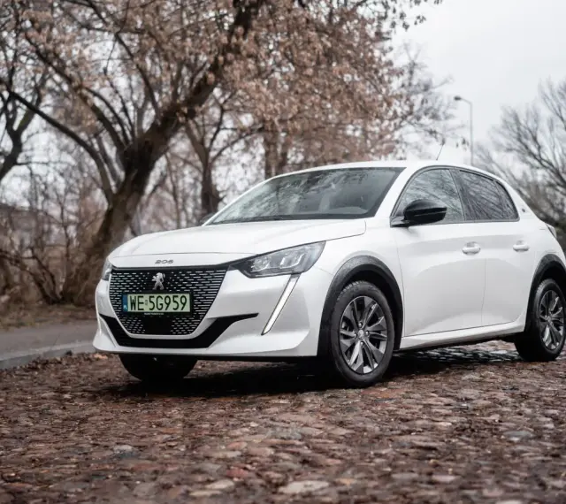 Biały Peugeot e-208 na brukowanej ulicy, z drzewami w tle.