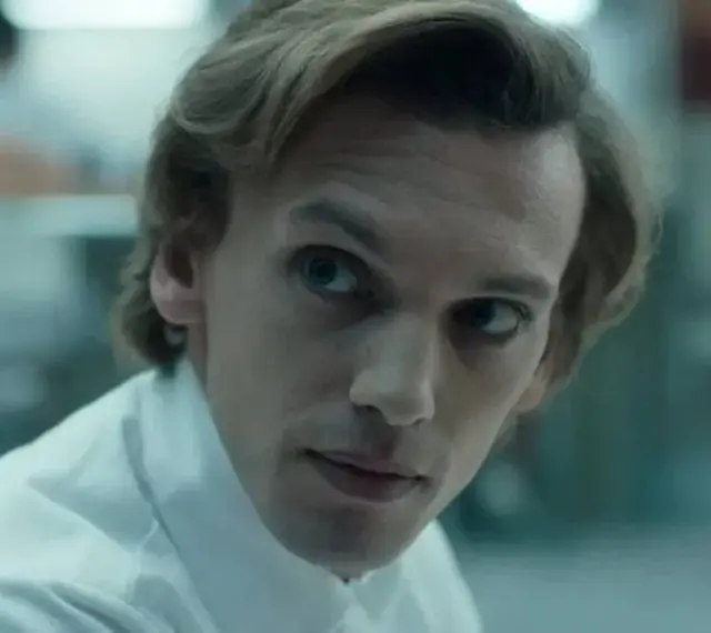 Henry Creel, znany jako Vecna z 001 stranger things, w białej koszuli, patrzy z uwagą.