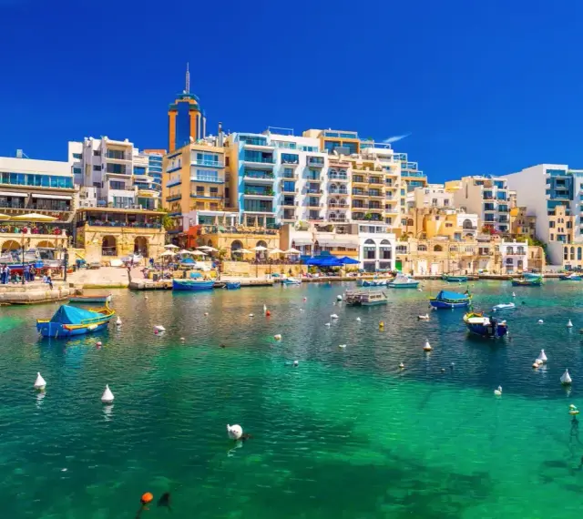 Malta, gdzie na Malcie jest tak pięknie! Woda krystalicznie czysta, łodzie kołyszą się na falach, a w tle kolorowe budynki.