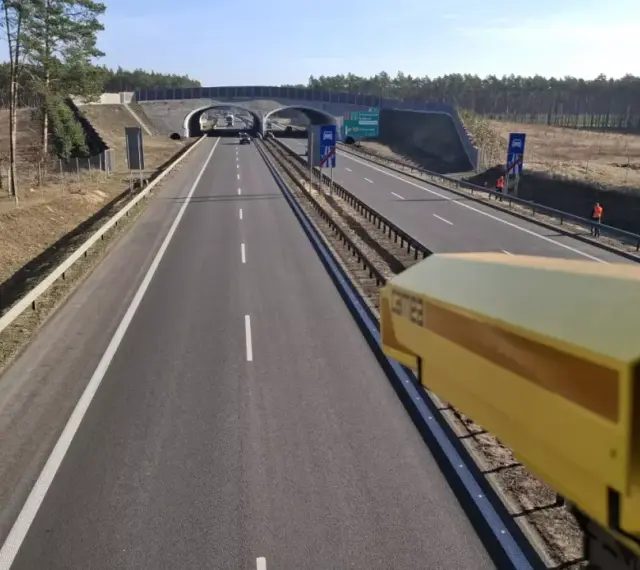 Żółta kamera monitoruje odcinek pomiarowy prędkości na S8. Droga prowadzi przez las, pod wiaduktem.
