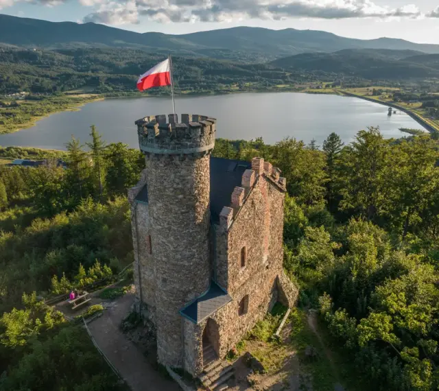 Kamienny zamek henryka z polską flagą na wieży, otoczony lasem, z jeziorem i górami w tle.