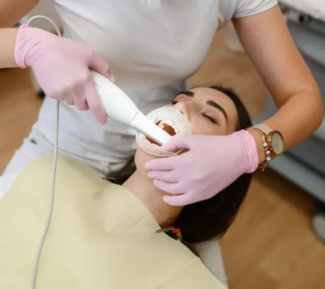 Dentystka w różowych rękawiczkach wykonuje zabieg piaskowania zębów. Czy piaskowanie zębów boli? Pacjentka ma otwarte usta i rozwierak.