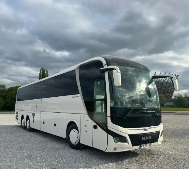 Biały MAN Lion's Coach, nowoczesny autokar turystyczny, zaparkowany na żwirowym placu pod pochmurnym niebem.