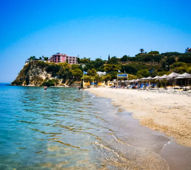 Piękne, turkusowe morze na Zakynthos. Plaża z leżakami i parasolami, a na klifie hotel.