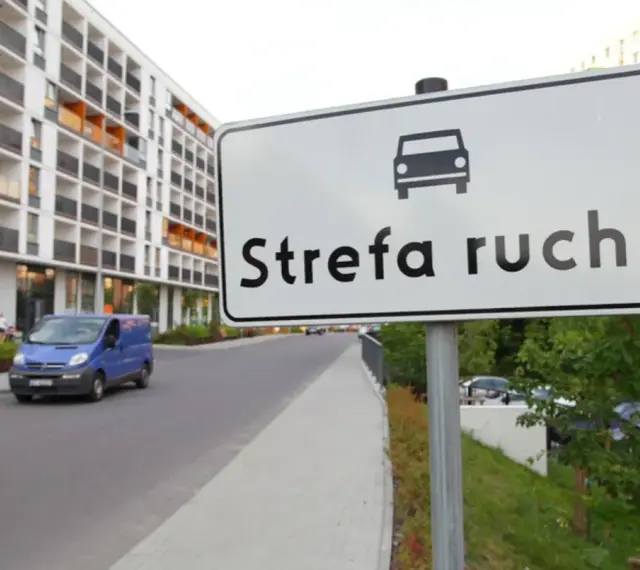 Znak "Strefa ruchu" przed nowoczesnym budynkiem mieszkalnym. Niebieski bus jedzie po drodze.