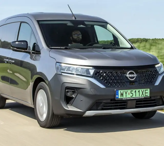 Szary Nissan Townstar jedzie po drodze, otoczony zielonymi polami. Kierowca w okularach prowadzi pojazd.