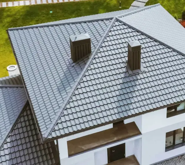Nowoczesny dom z szarym, czterospadowym dachem z blachodachówki. Dwa kominy wystają ponad linię dachu.