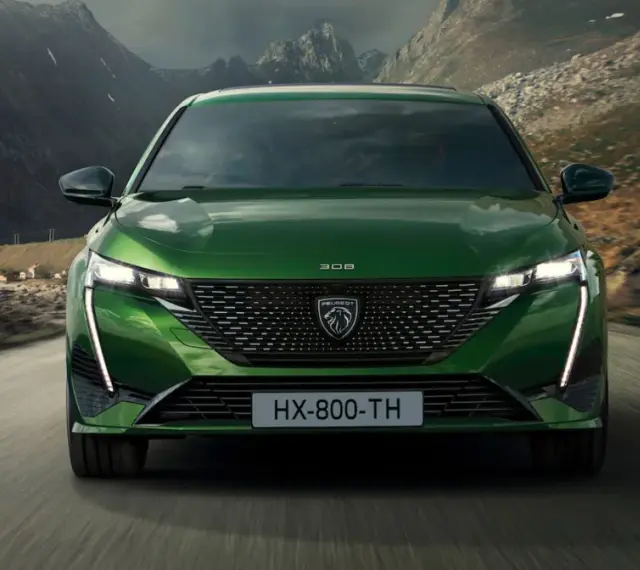 Zielony Peugeot 308 T7 jedzie drogą w malowniczym, górskim krajobrazie z widokiem na małą wioskę nad wodą.