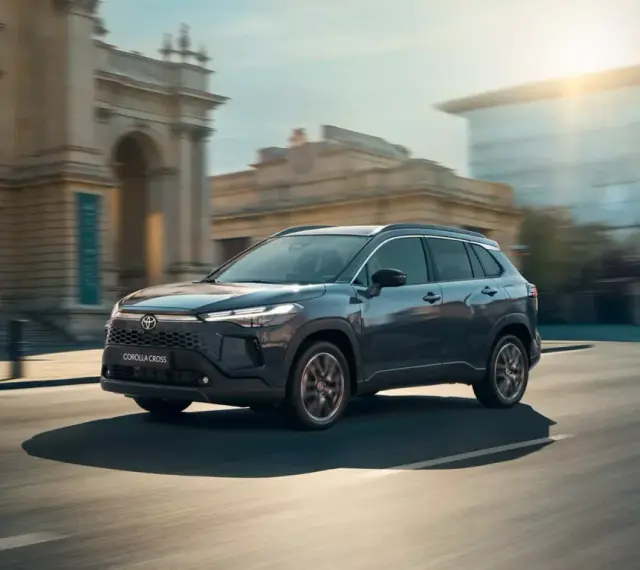 Ciemnoszary Toyota Corolla Cross Hybrid sunie ulicą miasta, odbijając światło słoneczne od nowoczesnych budynków.