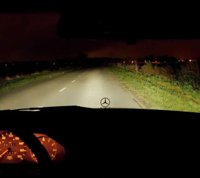 Widok z wnętrza samochodu nocą. Włączone światła drogowe oświetlają pustą drogę. Na desce rozdzielczej widać podświetlone zegary.