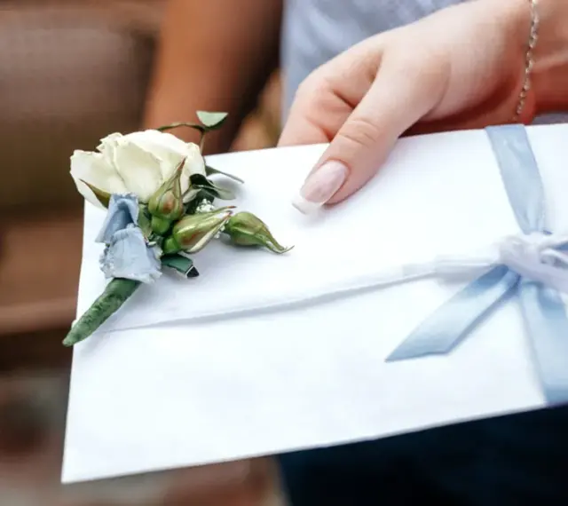 Dłoń trzyma białą kopertę z błękitną wstążką i ozdobnym kwiatkiem. Rozważasz skutki wręczenia pustej koperty lub zbyt małej kwoty? Opinie i etyka są tu kluczowe.