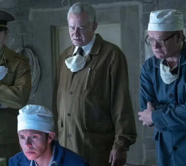 Mężczyźni w mundurach i fartuchach, w tym jeden w wojskowym, analizują coś na ekranie. Scena z serialu Czarnobyl Netflix.