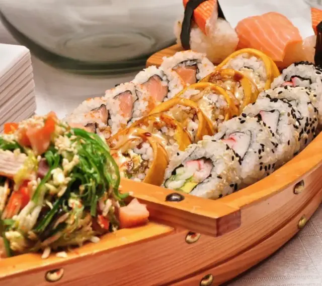 Drewniana łódka pełna sushi, dania z kraju, z którego pochodzi sushi, czyli Japonii. Obok stos białych talerzy.