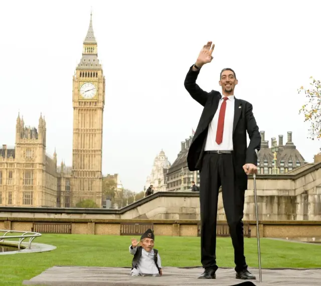 Der größte Mensch der Welt winkt vor dem Big Ben in London. Neben ihm steht ein Mann von sehr kleinem Wuchs.