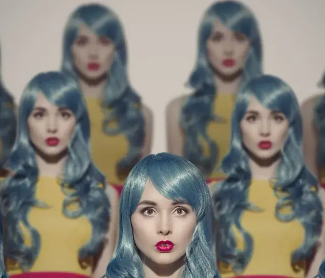 Mehrere Frauen mit blauen Perücken und roten Lippen scheinen sich zu klonen, eine surreale Szene.